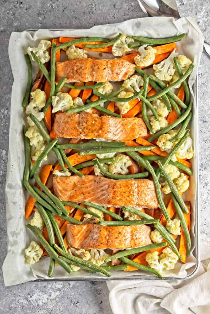 Cooked salmon and veggies sheet pan meal including green beans, cauliflower, and sweet potatoes displayed on the sheet pan. On a grey speckled background with a white cloth napkin and a large silver serving spoon poking into the frame.