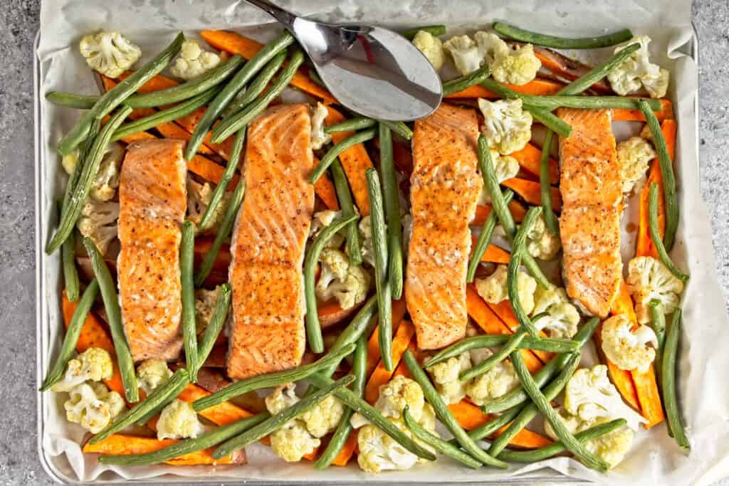 Cooked sheet pan salmon and vegetables meal including green beans, cauliflower, and sweet potatoes displayed on the sheet pan. On a grey speckled background with a large silver serving spoon resting on the baking sheet.