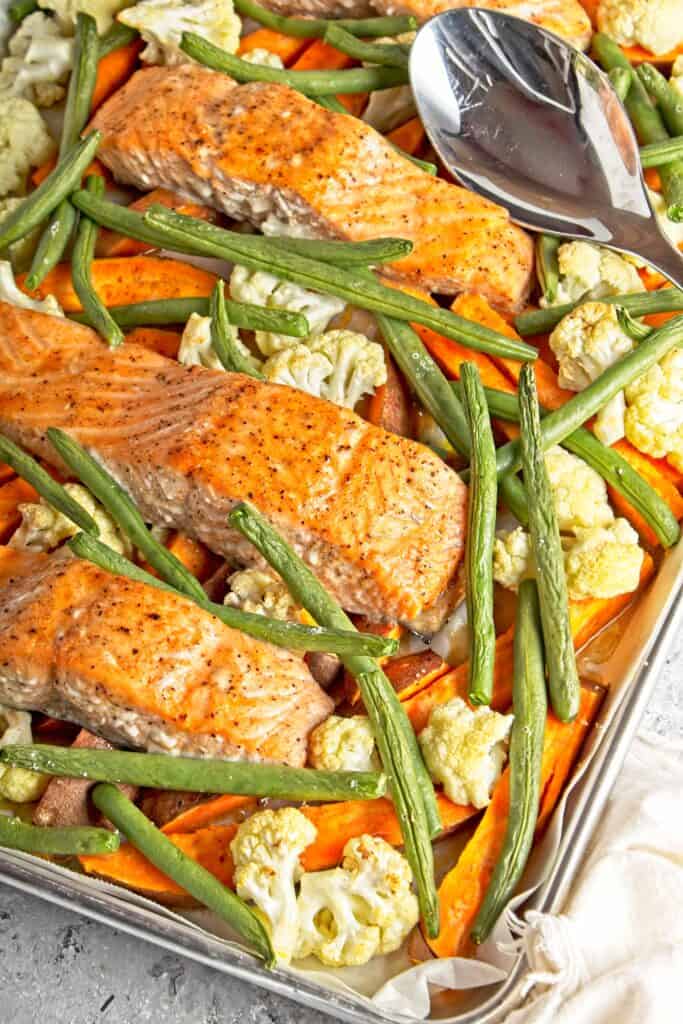Side view of cooked sheet pan salmon and vegetables meal including green beans, cauliflower, and sweet potatoes displayed on the sheet pan. On a grey speckled background with a large silver serving spoon resting on the baking sheet.