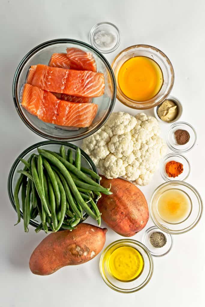 Ingredients for sheet pan salmon with vegetables in glass bowls on a white back ground. Salt, salmon filets, honey, mustard, cauliflower head, nutmeg, cayenne, whole green beans, whole sweet potatoes, vinegar, ground pepper, olive oil.