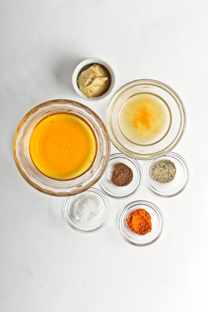 Ingredients for spicy honey glaze measured in 7 glass bowls of varying sizes: mustard, honey, vinegar, nutmeg, ground pepper, salt, cayenne. On a white background.