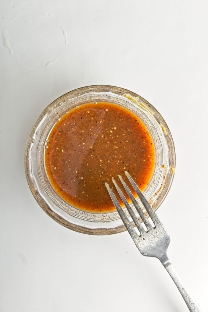 Small glass bowl with the whisked honey glaze and a fork perched on the edge of the bowl. On a white background.
