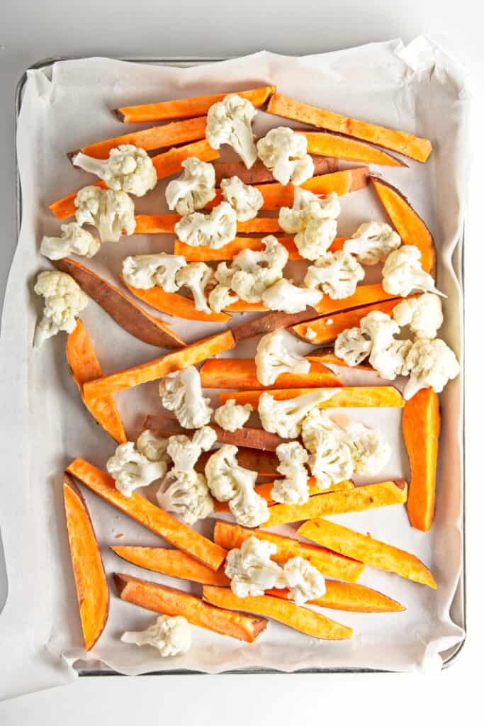 Sheet pan lined with baking parchment, scattered with slices of orange sweet potato and cauliflower florets. On a white background.
