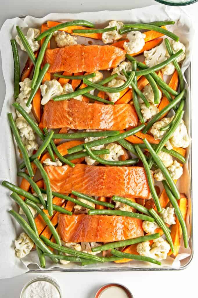 Fully assembled sheet pan meal with scattered sliced sweet potatoes and cauliflower florets, topped with glazed salmon filets and fresh green beans. Bowl of salt poking into the bottom of the frame.