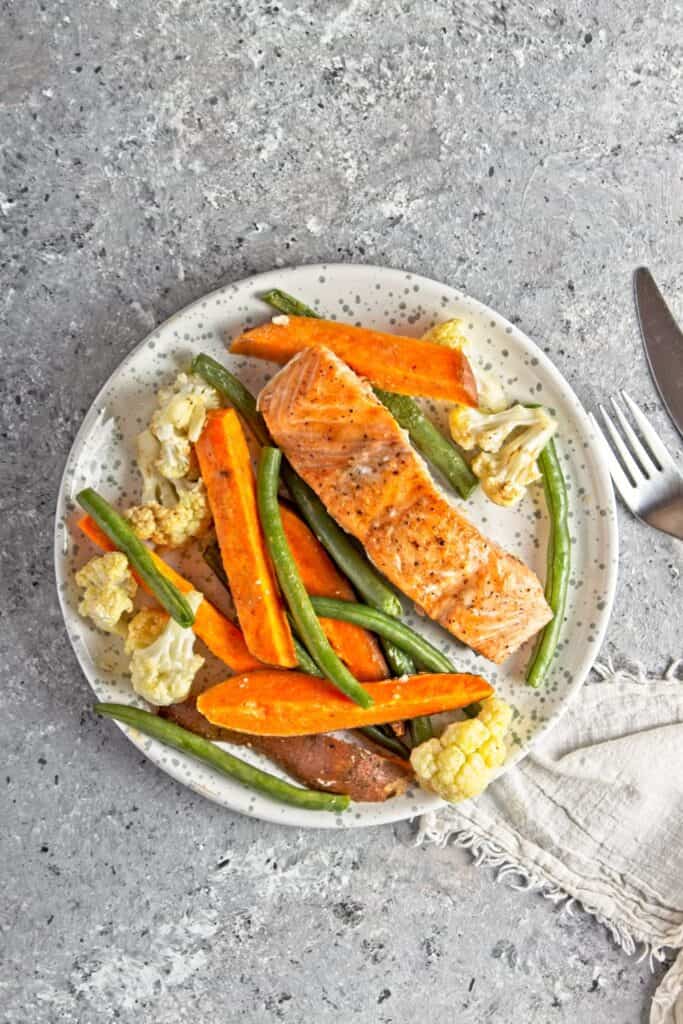 White and grey plate with a serving of sheet pan salmon and veggies on a grey speckled background. A white cloth napkin and a silver knife and fork to the right of the plate.